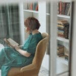 Kobieta czyta książkę w fotelu otoczonym regałami domowej biblioteki