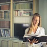 Kobieta w białej bluzce czytająca książkę w domowej bibliotece